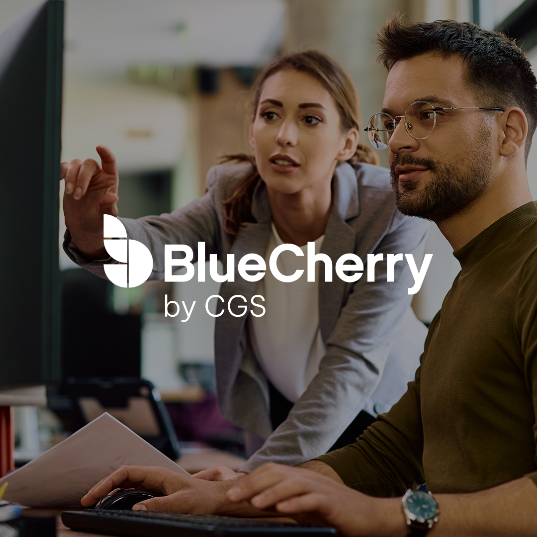 The BlueCherry logo overlayed on an image of two office workers at a desk, a man is sitting at the at the desk, while a woman is standing over him pointing at something being displayed on his computer monitor,.
