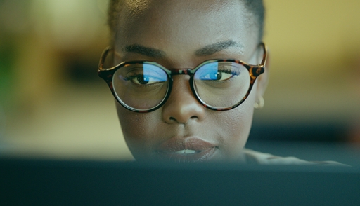 A woman working at a computer terminal, she is wearing glasses and the computer screen is reflected in the lenses.